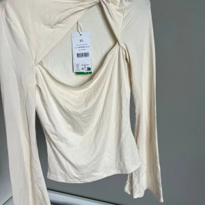 Offwhite twistad topp från NA-KD - Säljer en offwhite långärmad topp från NA-KD med twistad detalj framtill och draperad urringning. Tillverkad i mjukt, tunt material som känns skönt mot huden. Perfekt för dig som gillar stilrena och trendiga plagg.