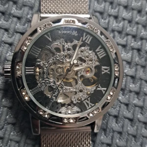 Silverfärgad mekanisk skelettklocka med mesh-armband - Stilren herrklocka i silver med synlig mekanism och romerska siffror på urtavlan. Klockan har ett mesh-armband i metall och glittrande stenar runt boetten. Perfekt för dig som gillar detaljer och mekanik.du kan ladda den utan batteri genom att snurra timvisaren en varv för att ladda den i ungefär 24timmar.helt ny oanvänd 
