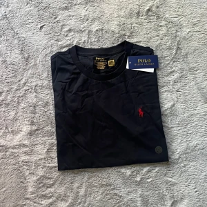 Svart t-shirt från Polo Ralph Lauren - Säljer en svart t-shirt från Polo Ralph Lauren med röd broderad logga på bröstet. Modellen är slim fit och har rund halsringning samt korta ärmar. Klassisk och stilren design med mjukt bomullsmaterial.