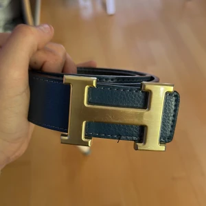  H-spänne - Säljer ett stilrent blått bälte med ett stort H-format spänne i guldfärg. Bältet är gjort i skinn och har en klassisk design som passar till många olika outfits.