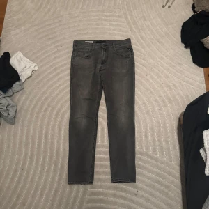 Grå Replay Anbass jeans - Säljer ett par grå jeans från Replay, modell Anbass. Byxorna har fem fickor, raka ben och klassisk knappgylf. Snygga detaljer och diskreta slitningar på baksidan ger en cool look. Perfekta för dig som gillar stilrena jeans med lite edge.
