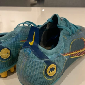 Nike Mercurial Vapor 14 Elite - Säljer ett par blå Nike fotbollsskor med gula och röda detaljer. Skorna har snörning och mönstrad ovansida, samt dobbar för spel på gräs. Swoosh-logga i rött och gult på sidan och M-logga bak. Perfekta för dig som vill sticka ut på planen!