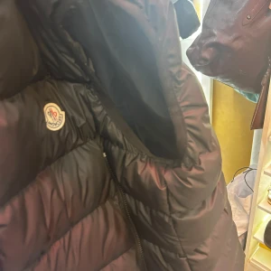 Svart väst från Moncler - Säljer en svart dunjacka från Moncler med klassisk logotyp på bröstet och dragkedja framtill. Jackan har en vadderad design och Moncler-tryck på insidan. Perfekt för kyliga dagar.