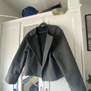Mörkgrå vårjacka från Gina Tricot - Säljer en mörkgrå croppad jacka från Gina Tricot. Jackan har en stilren design med krage och lång ärm. Perfekt för lager-på-lager och passar till många olika outfits.