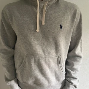 Grå hoodie från Ralph Lauren - Säljer en klassisk grå hoodie från Ralph Lauren med broderad logga på bröstet. Tröjan har huva med vita snören och en stor magficka. Perfekt för en avslappnad stil och enkel att matcha med det mesta.