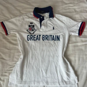 Vit piké från Polo Ralph Lauren med Great Britain-tryck - Vit pikétröja från Polo Ralph Lauren med blå och röda detaljer på krage och ärmslut. Stor text 'Great Britain' framtill, broderad logga och emblem med brittisk flagga. Klassisk pikékrage och korta ärmar.