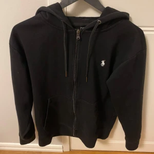 Svart zip hoodie rån Polo Ralph Lauren - Svart zip hoodie med dragkedja från Polo Ralph Lauren. Snygga men enkla fickor framtill och vit broderad logga på bröstet. Samt en luva, inga defekter på tröjan då den knappt är använd.