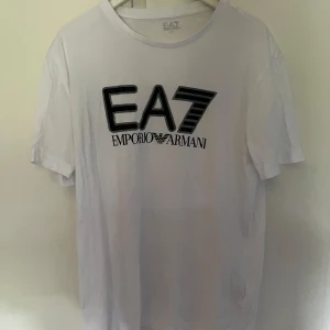 Vit EA7 Emporio Armani t-shirt - Väldigt fint skick. Vit t-shirt som är använd 4-5 gånger från EA7 Emporio Armani med stor svart logga framtill. Klassisk rund halsringning och korta ärmar. Perfekt för dig som gillar stilrena och trendiga märkesplagg. Storlek L men passar precis mig som har M i vanliga fall så passa folk med M och S