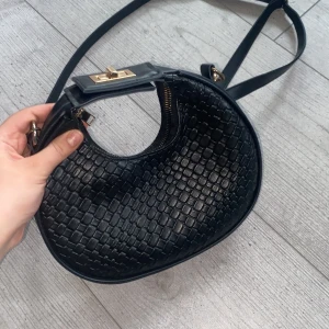 Svart axelväska i flätat skinn - Säljer en svart axelväska i rund form med flätad skinnstruktur och guldfärgade metalldetaljer. Väskan har ett snyggt vridlås och dragkedja upptill samt justerbar axelrem. Perfekt storlek för mobilen och andra saker.