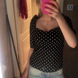 Svart prickig blus med puffärm - Supersöt svart blus med vita prickar och puffiga korta ärmar. Snygg sweetheart-urringning och figurnära passform som ger en trendig siluett. Perfekt att styla med jeans eller kjol för en cool look.