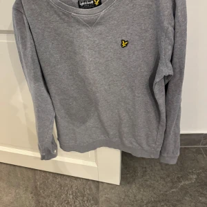 Grå sweatshirt från Lyle & Scott - En stilren grå sweatshirt från Lyle & Scott med klassisk rund hals och deras ikoniska gula fågel-logga broderad på bröstet. Tröjan har långa ärmar och är tillverkad i mjuk bomull, perfekt för en avslappnad och snygg look.