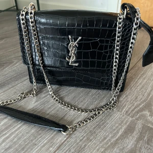 Svart axelväska från Saint Laurent - Säljer en svart axelväska från Saint Laurent i croco-präglat skinn med silverfärgad kedja och YSL-logga framtill. Väskan har ett elegant och stilrent utseende, perfekt för att lyfta vilken outfit som helst. Magnetlås och innerfack för smidig förvaring.