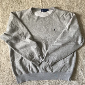 Grå sweatshirt från Polo Golf - Säljer en klassisk grå sweatshirt från Polo Golf med broderad logga på bröstet och Valley Brook-detalj på ärmen. Tröjan har rund halsringning och ribbade muddar vid ärmslut och nederkant. Perfekt för en avslappnad stil.