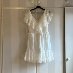 Vit volangklänning med v-ringning - Söt vit klänning med v-ringning och volangdetaljer upptill. Aldrig använd, prislappen finns kvar
