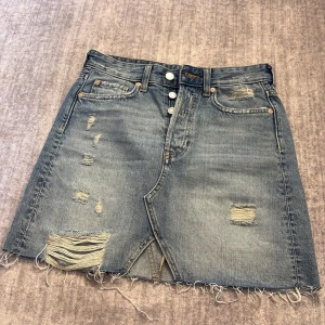 Blå jeanskjol från H&M - Säljer en jeanskjol från H&M med slitna detaljer och råa kanter. Kjolen har knappar framtill och klassiska fickor både fram och bak. Perfekt för dig som gillar en avslappnad och trendig stil.