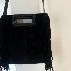 Svart Maje väska (stora modellen) - Säljer en svart axelväska från Maje i mocka med fransar längs sidorna och ett rektangulärt handtag upptill. Väskan har en avtagbar axelrem och stilren design som sticker ut. Använd fåtal gånger, så den är i ett väldigt finns skick🥰(Väskan är i den stora modellen).