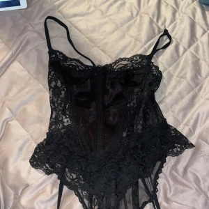 Svart spetskorsett med volang - Säljer en svart korsett- body med tunna axelband och vacker spets över hela plagget. Den har volangdetaljer nedtill och transparent mesh på sidorna och ryggen. Perfekt för att skapa en edgy och snygg look. 