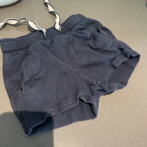 Mörkblå shorts från Newbie, strl 68 - Mörkblå shorts från Newbie i mjuk bomull med elastisk midja och snörning. Shortsen har två fickor fram och två bak, samt uppvikta benslut för en chill look. Perfekta för varma dagar. Tvätt påverkat men rena och hela 