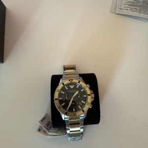 Emporio Armani guld och silverklocka - Nypris 5400 kr (489 euro), se sista bilden för bevis! Stilren set med klocka från Emporio Armani med svart urtavla, guldiga detaljer och silverfärgad länkarmband med guldfärgade inslag. Klockan har kronograf, datumvisning och tydliga index. Perfekt accessoar för dig som vill sticka ut med en lyxig look. Mått vid en enhetsstorlek: armbandsbredd 2cm, diameter på höljet 4,3cm, höjd på höljet 1,2cm. 