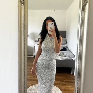 Glittrig silver maxiklänning - Säljer en lång, tight och glittrig silverklänning med hög hals och ärmlös design. Klänningen har öppen rygg och är täckt av paljetter som ger en riktigt festlig look. Perfekt för dig som vill sticka ut och glänsa på nästa bal eller fest.