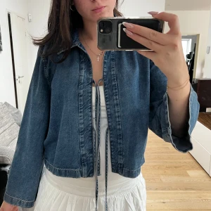 Blå jeansjacka med snörning - Säljer en blå jeansjacka med snygga snörningsdetaljer framtill. Jackan har en avslappnad passform, klassisk krage och vida ärmar. Andvänd 1 gång