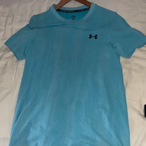 Blå träningst-shirt från Under Armour - Snygg ljusblå t-shirt från Under Armour med klassisk logga på bröstet. Perfekt för träning med sitt lätta och ventilerande syntetmaterial. Kortärmad modell med rund hals och sportig vibe.
