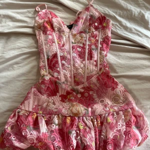 Rosa blommig kortklänning från Mistress Rocks - Supersöt kortklänning med blommigt mönster i rosa, vitt och inslag av gult. Klänningen har smala axelband, v-ringning och markerad midja med volang nertill. Materialet är lätt och känns som polyester, perfekt för sommarens alla tillfällen. Klänningen är från pretty little thing💕