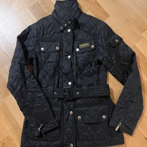 Svart quiltad jacka från Barbour - Quiltad svart jacka från Barbour International med bälte i midjan, hög krage och flera fickor med tryckknappar. Jackan är knappt använd & helt utan anmärkningar, så i nyskick!