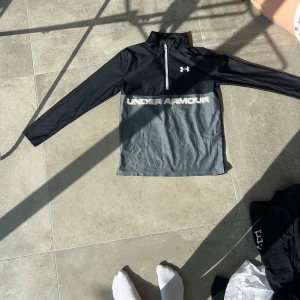 Svart och grå Under Armour half zip - Sportig långärmad tröja från Under Armour i svart och grått med stor logga och text framtill. Tröjan har half zip, hög krage och är tillverkad i ett lätt och ventilerande syntetmaterial, perfekt för träning. Loose fit för en avslappnad känsla.