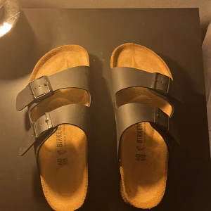 Svarta Birkenstock sandaler i skinn - Klassiska svarta Birkenstock sandaler med två breda remmar och justerbara spännen. Ovandel i skinn och innersula i mjukt material för extra komfort. Sula i slitstark gummi med tydligt mönster. Bara höra av er för fler bilder. Använda 1 gång helt nya