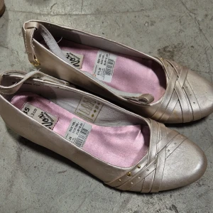 Ballerinaskor - Snygga ballerinaskor i beige med kilklack och rund tå. Skorna har dekorativa remmar med små guldfärgade nitar framtill och en justerbar rem runt hälen. Insidan är fodrad med ljusrosa tyg för extra stilpoäng.