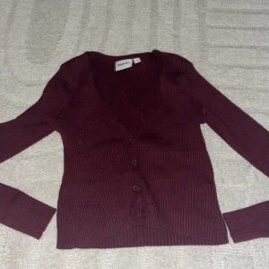 Vinröd ribbad kofta från Kappahl - Mysig vinröd kofta från Kappahl med ribbad struktur och v-ringning. Koftan har långa ärmar och knäpps med fyra knappar framtill. Perfekt att slänga över en topp för en chill och snygg look.