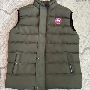 Canada Goose väst - Säljer en olivgrön dunväst från Canada Goose med klassisk logga på bröstet. Västen har ståkrage, två fickor med tryckknappar och quiltad design. Perfekt för lager-på-lager och snygg till streetwear. Fodret är svart och glansigt.