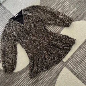 Leopardmönstrad kortklänning med volang - Säljer en kort klänning med leopardmönster och volang nertill. Klänningen har långärmade puffärmar, markerad midja och v-ringad hals. Färgerna går i svart, beige och brunt. Perfekt för dig som vill sticka ut med en trendig look.