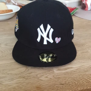 New Era Yankees 59FIFTY keps svart - Svart New Era 59FIFTY keps med New York Yankees-logga i vitt framtill, broderat hjärta med baseballsömmar, World Series-märke på sidan och texten 'Love & Hustle = Game' broderad bak. Platt skärm och klassisk passform. Insidan har märkets band och etiketter. Säljer pga har ingen användning till det.