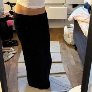 Svart långkjol i mjukt tyg - Säljer en svart långkjol med rak passform och låg midja. Kjolen är enkel och stilren, perfekt om du gillar minimalistisk stil. Materialet ser ut att vara mjukt och följsamt, vilket gör den riktigt skön att bära.