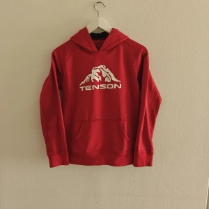 Tendon hoodie  - Röd hoodie från Tenson med vit logga och bergmotiv på bröstet. Klassisk känguruficka framtill, huva och långa ärmar. Mjuk insida och skön passform, perfekt för chill dagar eller när du vill ha en sportig look.