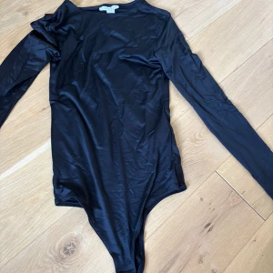 Svart långärmad bodysuit från Missguided - Svart, figurnära bodysuit från Missguided med långa ärmar och rund hals. Tillverkad i mjukt, stretchigt material med tryckknappar i grenen för enkel på- och avtagning. Perfekt att styla med jeans eller kjol för en clean look.