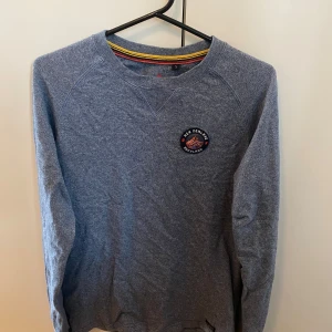 Blå sweatshirt från NZA Auckland - Blåmelerad sweatshirt från NZA New Zealand Auckland med rund hals och broderad patch på bröstet. Tröjan har långa ärmar och klassisk passform, perfekt för en chill och stilren look. Mjuk bomullskänsla och snygga detaljer vid halsringningen. Ny pris 2 tusen