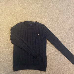 Mörkblå kabelstickad tröja från GANT - Säljer en mörkblå kabelstickad tröja från GANT i premium bomull. Tröjan har rund halsringning, långa ärmar och GANT-logga broderad på bröstet. Perfekt för dig som gillar klassisk och stilren look. Priset är diskuterbart!