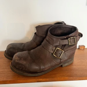 Bruna boots i skinn med spännen - Tuffa bruna boots i äkta skinn med två metallspännen på varje sida och stålsätta inuti. Kraftig sula och rund tå ger en robust känsla. I använt skick men går säkert att snygga till för den som kan och orkar. Se bilder för slitage på skorna.