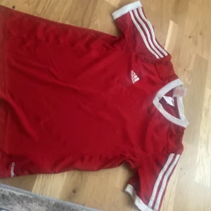 Röd Adidas fotbollströja med vita detaljer - Röd fotbollströja från Adidas med klassiska vita ränder på axlarna och vita muddar vid ärmslut och hals. Tillverkad i lätt och ventilerande material, perfekt för träning eller match. Adidas-logga på bröstet och enkel, sportig design. Ligger i Vetlanda.