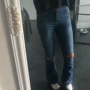 Blå bootcut jeans med slitningar - Snygga blå jeans med bootcut-modell och slitningar vid knäna. Jeansen har hög midja och klassisk femficksdesign. Perfekta för dig som gillar en avslappnad men trendig look.