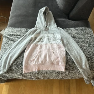 Grå och rosa hoodie med New York-tryck H&M - Snygg hoodie från H&M i grått och ljusrosa med stort 'NEW YORK'-tryck framtill. Tröjan har huva, lång ärm och en  magficka.Använd några gånger.  