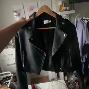 Svart croppad skinnjacka Vero Moda - Snygg svart croppad skinnjacka från NA-KD med klassisk bikerstil, silvriga dragkedjor och knappar. Jackan har långa ärmar och coola detaljer som ger en edgy vibe. Perfekt att slänga över en hoodie eller t-shirt för en grym look.