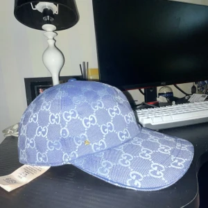 Blå Gucci keps med GG-mönster - Snygg blå keps från Gucci med ikoniskt GG-mönster över hela kepsen. Tillverkad i jeansliknande bomullsmaterial med justerbar rem bak. Perfekt för dig som vill sticka ut med en lyxig och trendig accessoar. Helt ny är kepsen. De är bara och höra av er om frågor och så
