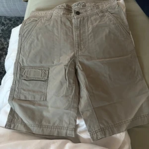 Beige cargo shorts från Sail Racing - Snygga beige cargo shorts från Sail Racing med flera praktiska fickor, bland annat en med lock på sidan. Klassisk passform och tillverkade i bomull, perfekta för varma dagar. Broderad logga baktill och bälteshällor för extra stil.