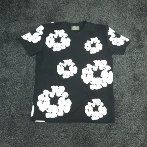Svart t-shirt med blommor från Denim Tears - Svart t-shirt från Denim Tears med stora vita blommiga tryck över hela plagget. Klassisk passform med rund hals och korta ärmar. Perfekt statement-piece för dig som vill sticka ut med en unik och trendig look.