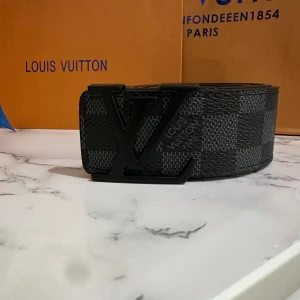  rutigt svart bälte - Snyggt svart bälte med klassiskt rutigt mönster och stor Märkes-logga i matt svart metall. Bältet är gjort i exklusivt skinn och har en bred, modern design som sticker ut. Perfekt accessoar för att lyfta din outfit med en lyxig touch.