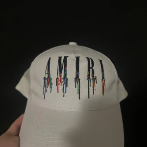 Amiri Keps One Size - Snygg vit keps från Amiri med broderad logga framtill där bokstäverna har coola färgstänk som rinner ner. Baksidan har meshpanel och justerbar snapback. Perfekt för dig som vill sticka ut med en unik accessoar.